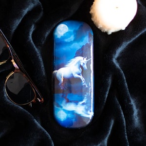 May include: A blue and white glasses case featuring a unicorn running through a moonlit sea. The case has a reflective surface.