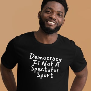 Può includere: T-shirt nero con la scritta bianca "Democracy Is Not A Spectator Sport". La maglietta è realizzata in un materiale morbido e ha maniche corte. La persona che indossa la maglietta sta sorridendo.