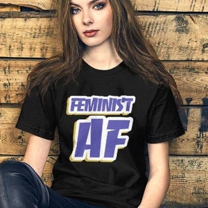 Puede incluir: Camiseta negra con el texto "FEMINIST AF" en letras moradas y amarillas. La modelo lleva vaqueros azul oscuro y está sentada junto a un casco rojo y blanco.