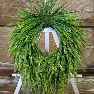 May include: A lush green artificial fern wreath, hanging from a white easel. The wreath is made of realistic-looking fern fronds, with a full and cascading shape.