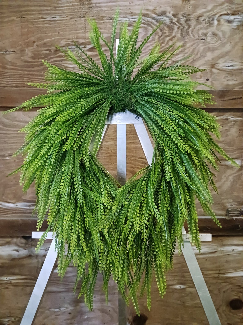 May include: A lush, cascading artificial greenery wreath, boasting a vibrant green hue. The wreath features numerous small, densely packed leaves, creating a full and textured appearance. It's displayed on a simple white easel, showcasing its shape and density effectively. The artificial plant is ideal for indoor or outdoor decoration, adding a touch of nature to any space. This faux greenery wreath is perfect for adding a pop of color and texture to your home decor.