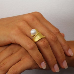 Könnte beinhalten: Ein goldener Ring mit einem weißen Perlmutt, das in einem gebogenen Band eingefasst ist. Der Ring wird an einem Finger getragen.