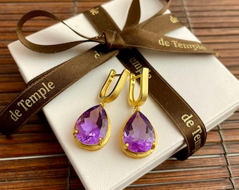 May include: A pair of gold earrings with purple teardrop-shaped gemstones. The earrings are in a white box with a brown ribbon that says "de Temple".