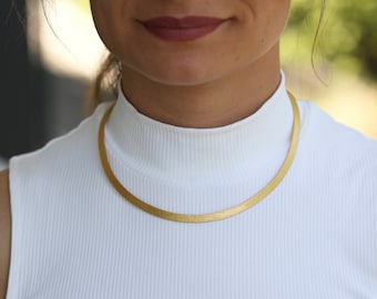 May include: A gold chain necklace with a flat, smooth surface. The chain is worn by a person with a white shirt.
