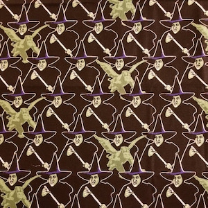 May include: A brown fabric with a repeating pattern of a witch with a green flying monkey. The witch is wearing a purple hat and a black robe. She is holding a broom in her right hand.