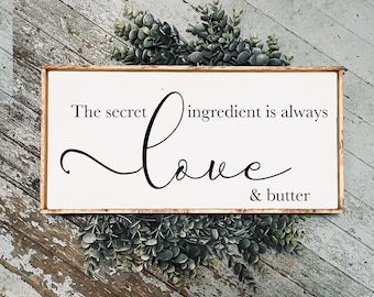 May include: A rectangular wooden sign with the text "The secret ingredient is always Love & butter" in black script. The sign has a white background and a brown wooden frame. Greenery is arranged around the sign.