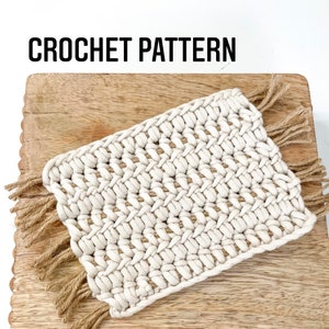 Puede incluir: Un posavasos de crochet blanco con flecos marrones sobre una superficie de madera con un borde de cuentas. El posavasos tiene un patrón tejido y está hecho de fibras naturales. El texto "CROCHET PATTERN" es visible en la parte superior de la imagen.