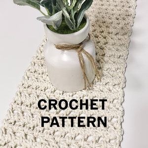 May include: A crocheted table runner in a cream colour with a simple, geometric pattern. The runner is displayed with a white vase and a green plant.