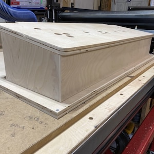 May include: A wooden box with a hinged lid, sitting on a plywood surface. The box has a series of holes drilled into the top.