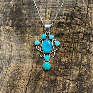 Puede incluir: Un collar con colgante de plata que presenta una gran piedra turquesa ovalada central rodeada por cuatro piedras turquesas redondas más pequeñas. El engaste de plata tiene un diseño decorativo de alambre en espiral y cuelga de una fina cadena.