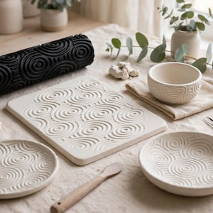 Könnte beinhalten: Eine schwarze zylindrische Tonrolle mit Spiralmuster, eine rechteckige Tonplatte, zwei runde Teller und eine kleine Schale, alle mit passenden geprägten konzentrischen Kreisdesigns, begleitet von einem kleinen Holzwerkzeug und einem Metallwerkzeug.