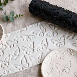 Könnte beinhalten: Eine schwarze zylindrische Musterwalze mit Schmetterlingsmotiv, platziert über einer rechteckigen weißen Tonplatte und zwei runden weißen Tonschalen, die alle mit einem sich wiederholenden Schmetterlingsdesign auf ihren Oberflächen geprägt sind.