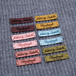 May include: A collection of rectangular labels in various colors, including brown, pink, and blue, with the text "Knitted by Isabella" and "Handmade by Olivia" in a cursive font. The labels are on a gray knitted fabric.