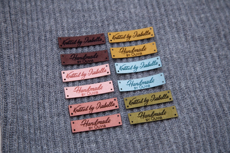 May include: A collection of rectangular labels in various colors, including brown, pink, and blue, with the text "Knitted by Isabella" and "Handmade by Olivia" in a cursive font. The labels are on a gray knitted fabric.