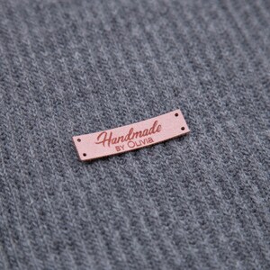 May include: A small, rectangular, pink leather tag with the words "Handmade by Olivia" in a cursive font. The tag has two small holes on each end and is set against a gray, ribbed fabric background.