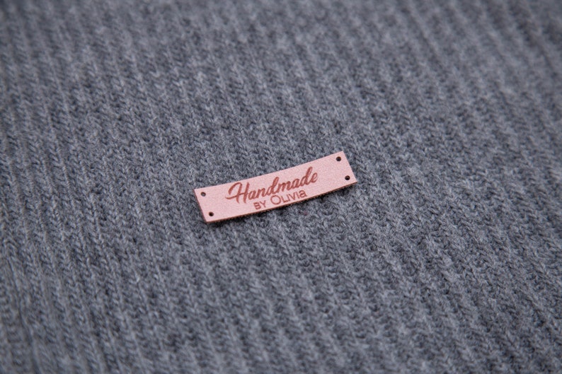May include: A small, rectangular, pink leather tag with the words "Handmade by Olivia" in a cursive font. The tag has two small holes on each end and is set against a gray, ribbed fabric background.