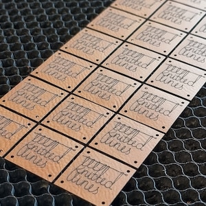 May include: Close-up of multiple square leather labels with the text "Knitted Lines" in a handwritten font. Each label has small holes in the corners. The labels are light brown and arranged on a dark surface.