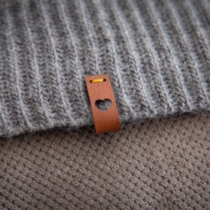 May include: A grey and brown knitted fabric with a brown leather tag featuring a heart cutout. The tag has a yellow stitch around the edge.