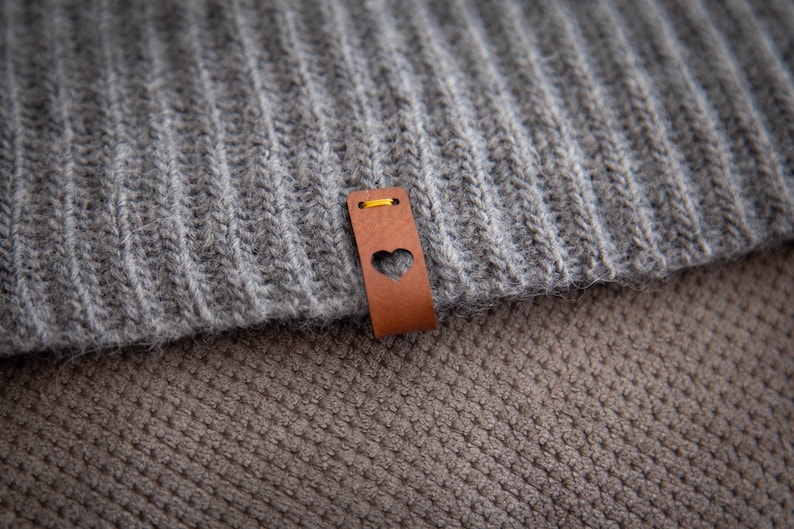May include: A grey and brown knitted fabric with a brown leather tag featuring a heart cutout. The tag has a yellow stitch around the edge.