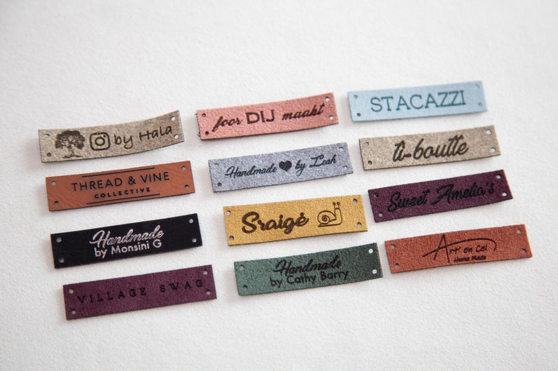 May include: A collection of 11 different leather labels with various text and designs. The labels are in different colors, including brown, beige, gray, blue, green, and red. The text on the labels includes "Thread & Vine Collective", "Handmade by Monsini G", "Sraigė", "Sweet Amelia's", "Village Swag", "Handmade by Cathy Barry", "O by Hala", "foor DIJ maakt", "STACAZZI", "U-boulte", and "Art on Col".