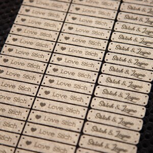 May include: Close-up of numerous rectangular wooden tags. Some tags read "Love Stich" with a heart symbol, while others say "Salah & Jayne." The tags have small holes at each corner, and are arranged in neat rows.
