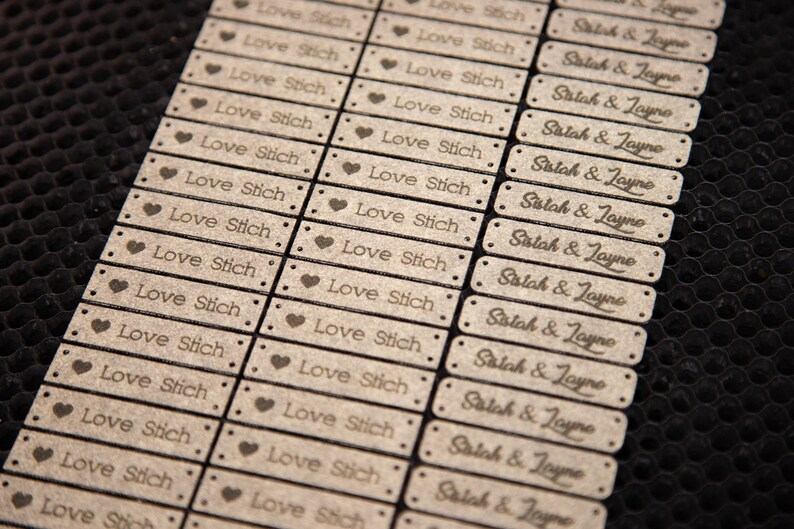 May include: Close-up of numerous rectangular wooden tags. Some tags read "Love Stich" with a heart symbol, while others say "Salah & Jayne." The tags have small holes at each corner, and are arranged in neat rows.