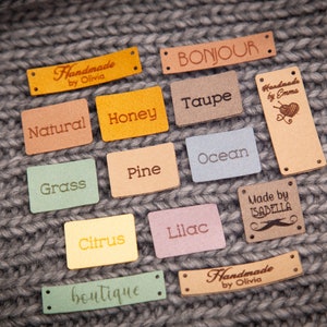 May include: A collection of 13 colorful leather labels with various text, including "Handmade by Olivia", "Bonjour", "Natural", "Honey", "Taupe", "Handmade by Emma", "Grass", "Pine", "Ocean", "Citrus", "Lilac", "Made by Isabella", and "boutique".