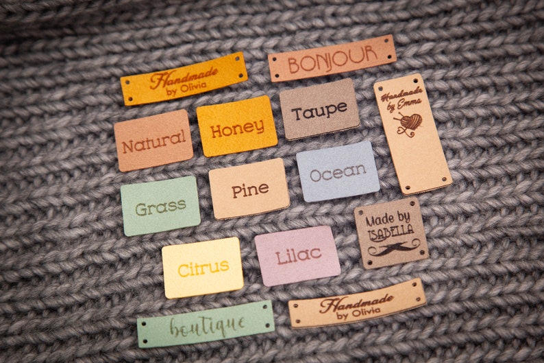 May include: A collection of 13 colorful leather labels with various text, including "Handmade by Olivia", "Bonjour", "Natural", "Honey", "Taupe", "Handmade by Emma", "Grass", "Pine", "Ocean", "Citrus", "Lilac", "Made by Isabella", and "boutique".