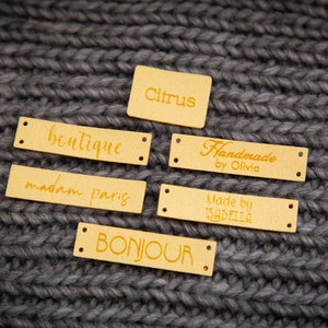 May include: Six gold leather labels with different text on each. The labels are on a gray knitted fabric background. The text on the labels reads: "boutique", "madam paris", "bonjour", "citrus", "Handmade by Olivia", and "Made by Isabella".