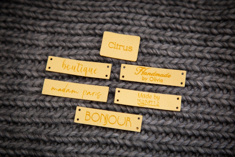 May include: Six gold leather labels with different text on each. The labels are on a gray knitted fabric background. The text on the labels reads: "boutique", "madam paris", "bonjour", "citrus", "Handmade by Olivia", and "Made by Isabella".