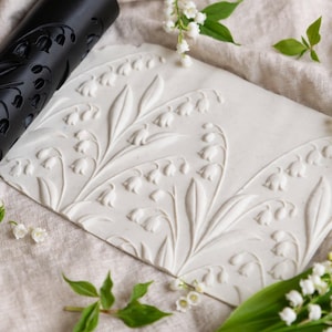 Puede incluir: Un rodillo cilíndrico negro con un patrón grabado de lirios de los valles y una placa rectangular de arcilla blanca con el mismo diseño floral en relieve, rodeados de tallos verdes frescos de lirios de los valles y flores blancas.