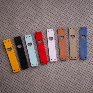 May include: Ten leather straps with a heart-shaped cutout in various colours, including yellow, black, light blue, red, white, brown, beige, and blue.