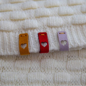 May include: Three fabric tags with heart cutouts sewn onto a white knitted fabric. The tags are yellow, red, and purple.