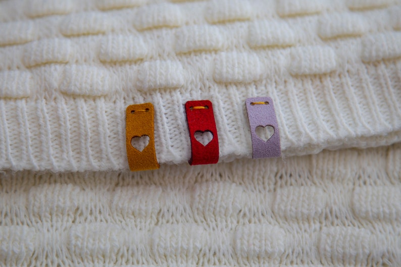 May include: Three fabric tags with heart cutouts sewn onto a white knitted fabric. The tags are yellow, red, and purple.