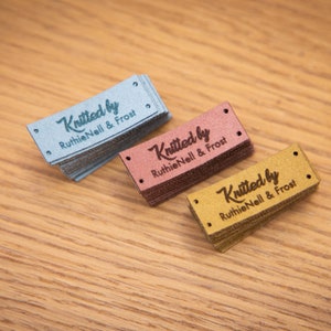 May include: Three stacks of rectangular labels in blue, rose, and gold. Each label is embossed with the words "Knitted by Ruthie Nell & Frost" in a script font. The labels have small holes at each end.