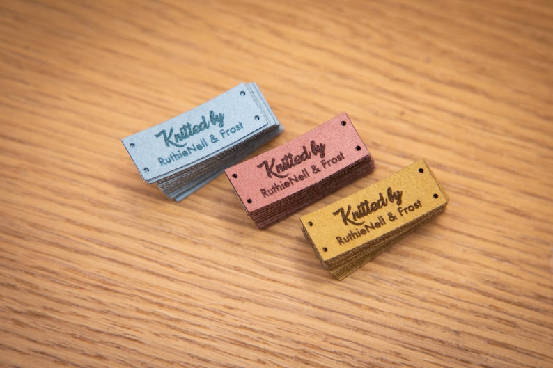 May include: Three stacks of rectangular labels in blue, rose, and gold. Each label is embossed with the words "Knitted by Ruthie Nell & Frost" in a script font. The labels have small holes at each end.