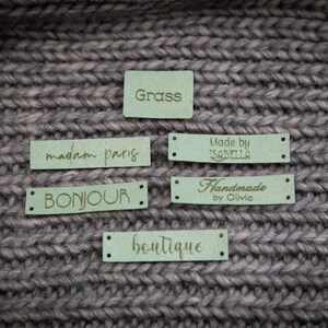 May include: Five green fabric labels with gold lettering. The labels read "Grass", "madam paris", "BONJOUR", "Handmade by Olivia", and "boutique".
