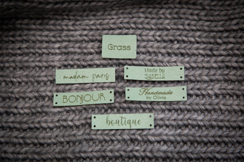 May include: Five green fabric labels with gold lettering. The labels read "Grass", "madam paris", "BONJOUR", "Handmade by Olivia", and "boutique".