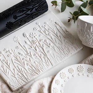 Puede incluir: Un rodillo cilíndrico negro de arcilla con un patrón de flores silvestres grabado, una placa rectangular de arcilla blanca con un diseño floral en relieve, un cuenco de cerámica blanca con relieve floral y un plato blanco con borde floral.