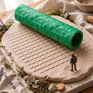 Könnte beinhalten: Eine leuchtend grüne, zylindrische Ton-Texturwalze mit Gittermuster, die auf einem flachen Stück hellbeigem Ton liegt. Eine kleine Miniaturfigur einer Person mit Rucksack steht auf der Tonoberfläche, die ein eingeprägtes Gitterdesign aufweist.