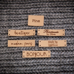 May include: Five brown leather labels with different text on each. The labels are on a gray knitted fabric background. The text on the labels reads: "Pine", "boutique", "Handmade by Olivia", "madam paris", "Made by ISABELLA", and "BONJOUR".