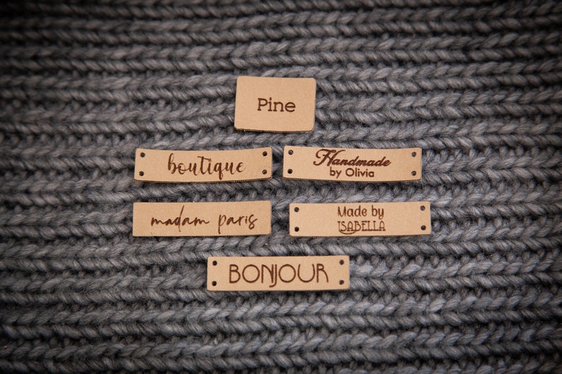 May include: Five brown leather labels with different text on each. The labels are on a gray knitted fabric background. The text on the labels reads: "Pine", "boutique", "Handmade by Olivia", "madam paris", "Made by ISABELLA", and "BONJOUR".