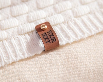 May include: Close-up of a cream-coloured knitted jumper with a textured pattern. A brown leather tag with the text "YOUR TEXT" is attached with a gold-coloured stud. The jumper has a ribbed hem.
