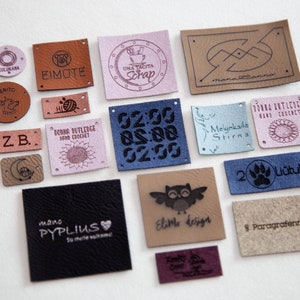 May include: A collection of 15 leather labels with various designs and text. The labels are in different colors, including brown, pink, blue, and beige. Some of the labels feature text, such as "Eimute", "Una Tacita Scrap", "Melnkaile Stirns", and "2 Li&uuml;tukai".