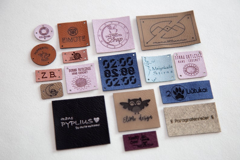 May include: A collection of 15 leather labels with various designs and text. The labels are in different colors, including brown, pink, blue, and beige. Some of the labels feature text, such as "Eimute", "Una Tacita Scrap", "Melnkaile Stirns", and "2 Li&uuml;tukai".