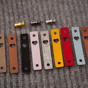 May include: Ten leather straps with a heart-shaped cutout, in various colors including brown, black, pink, gray, yellow, red, and blue. The straps are arranged in a row on a gray knitted fabric.