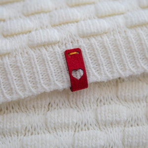 May include: White knitted fabric with a red heart-shaped tag attached. The tag has a gold thread border and a white heart cutout.