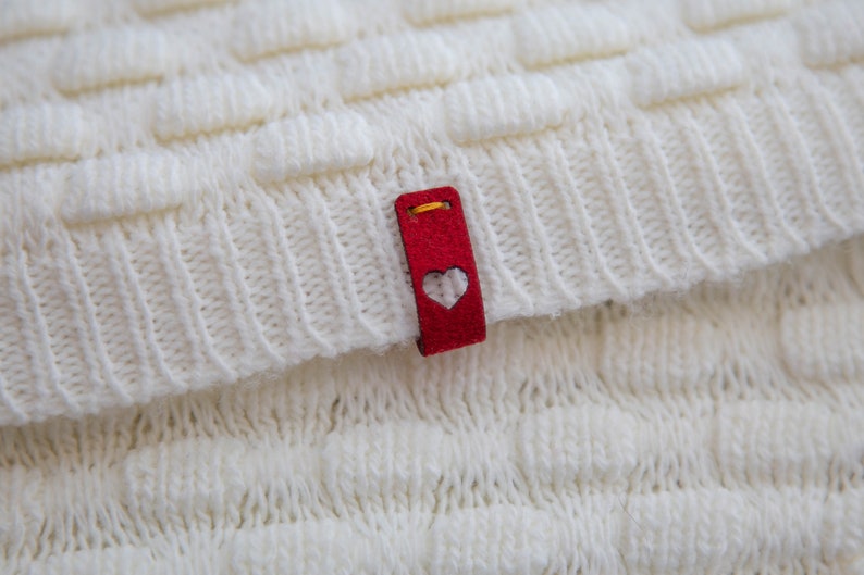 May include: White knitted fabric with a red heart-shaped tag attached. The tag has a gold thread border and a white heart cutout.