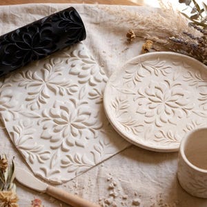 Op de afbeelding: Een zwarte cilindrische deegroller met een gebloemd reliëfpatroon, een platte plaat van witte klei met een bijpassende bloemenafdruk, een rond bord van witte klei met een bloemmotief en een witte keramische mok met een bijpassend bloemmotief.