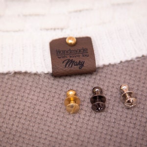 May include: A brown leather tag with the text "Handmade with love by Mary" and a gold rivet. Three metal fasteners, one gold and two silver, are shown below the tag.
